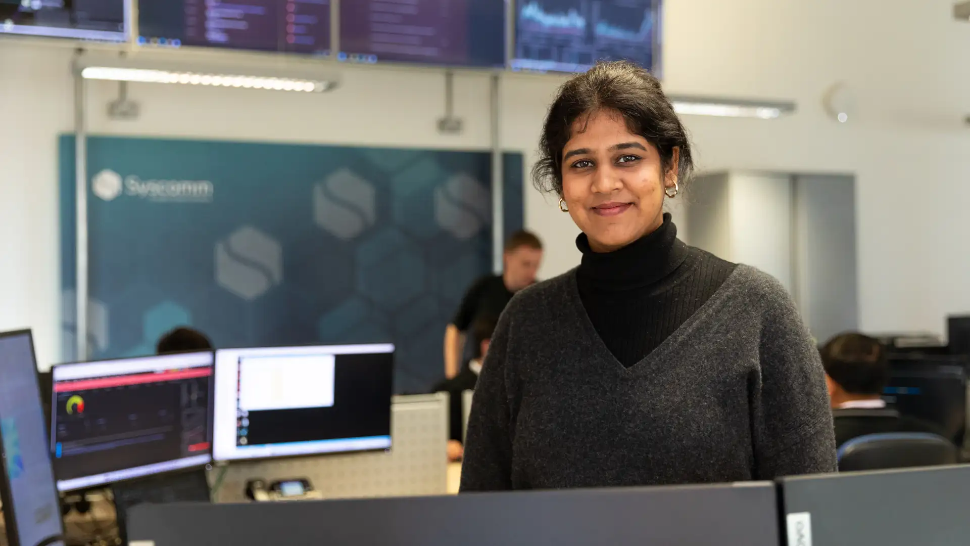 A confident woman in a grey sweater standing in front of multiple workstations and monitoring screens