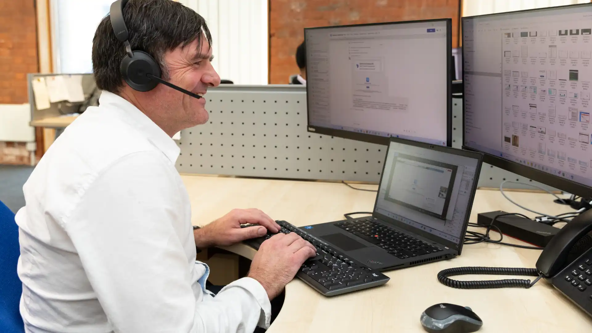 A man wearing a headset smiling while working on a laptop with multiple monitors displaying IT