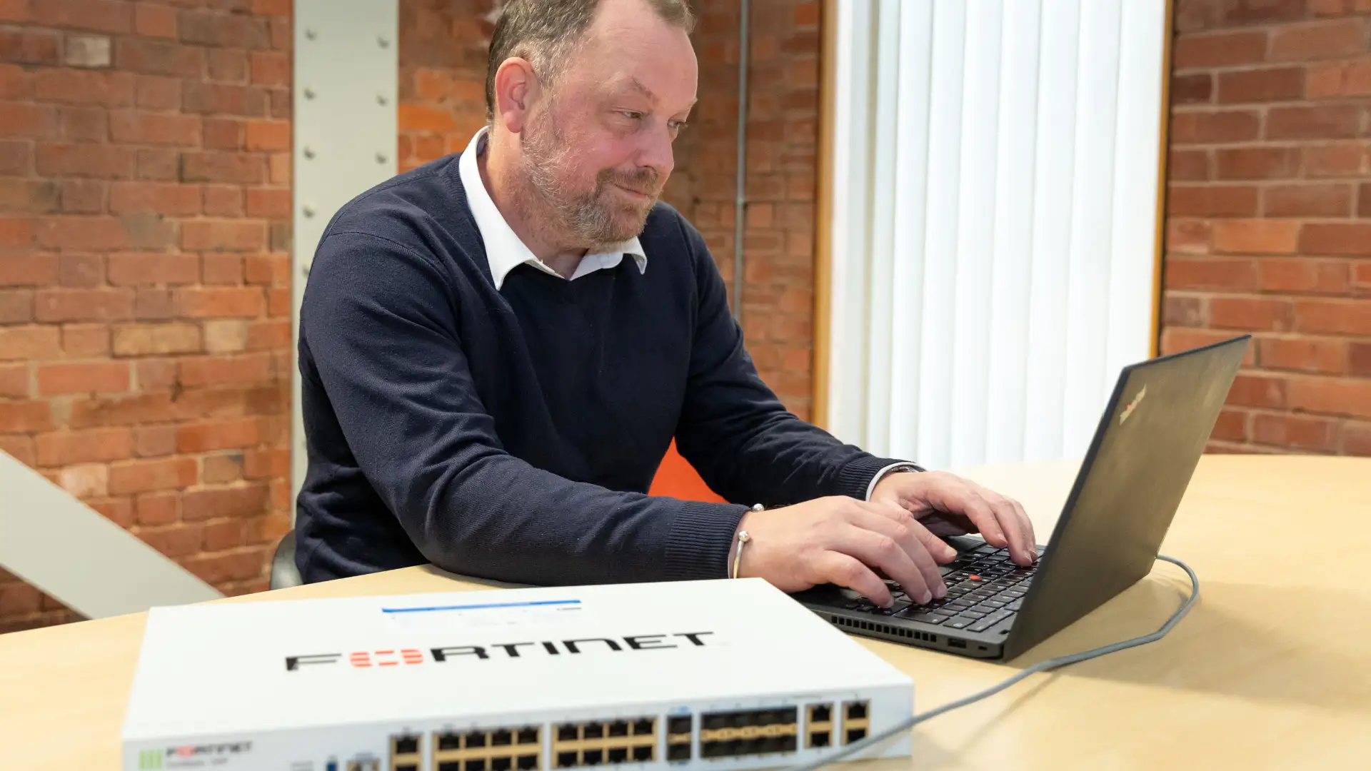 IT professional configuring a network device on a laptop with a Fortinet switch on the table in a modern office